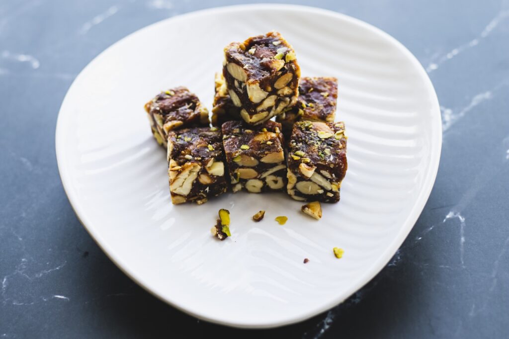 Stack of sugar-free nut and fruit dessert bars on a white ribbed plate, featuring pistachios and almonds, arranged on a dark slate surface